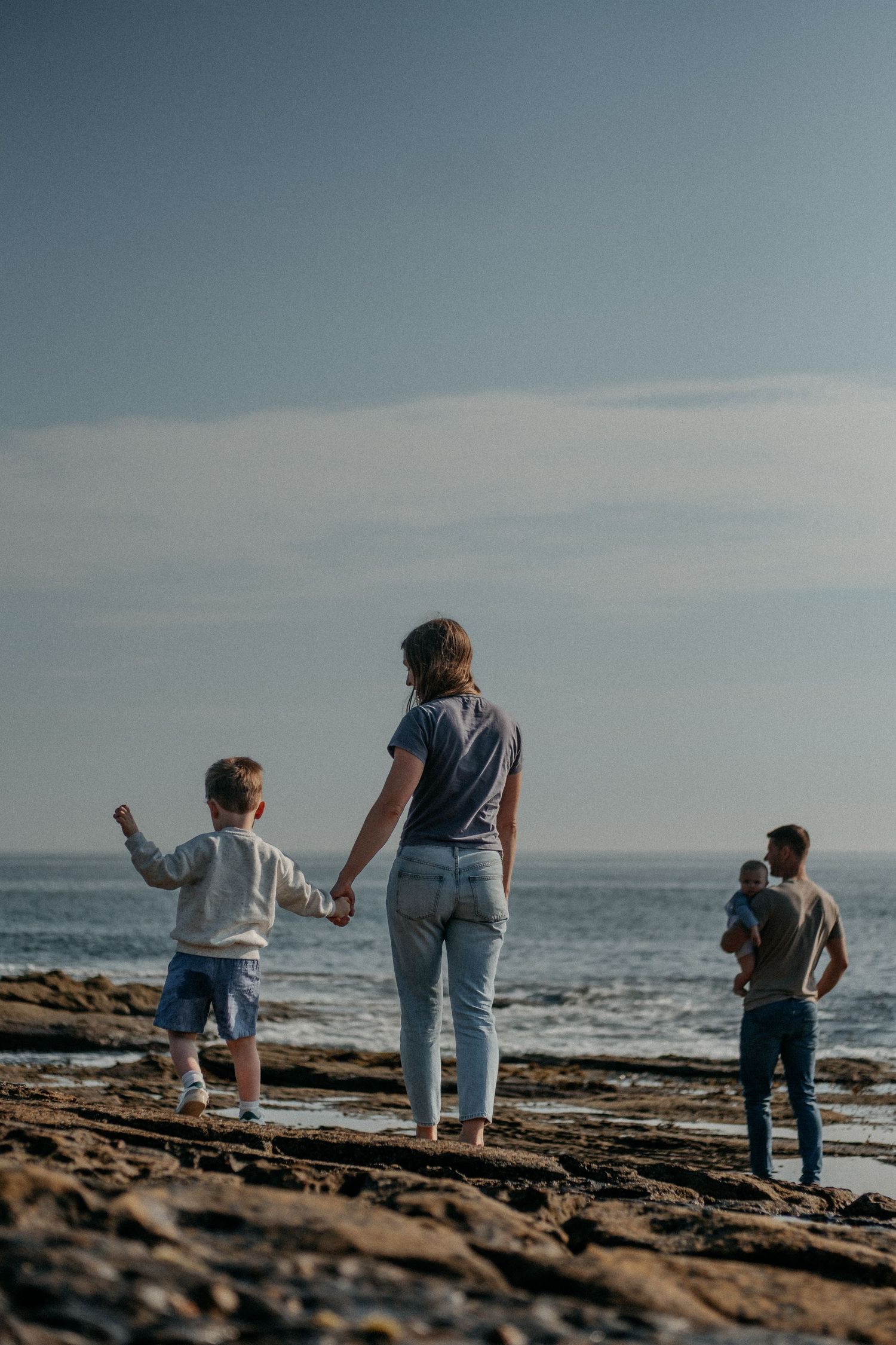 Family with young child standing on a beach watching waves and distant pier during a relaxed lifestyle family photography session in Alnwick
