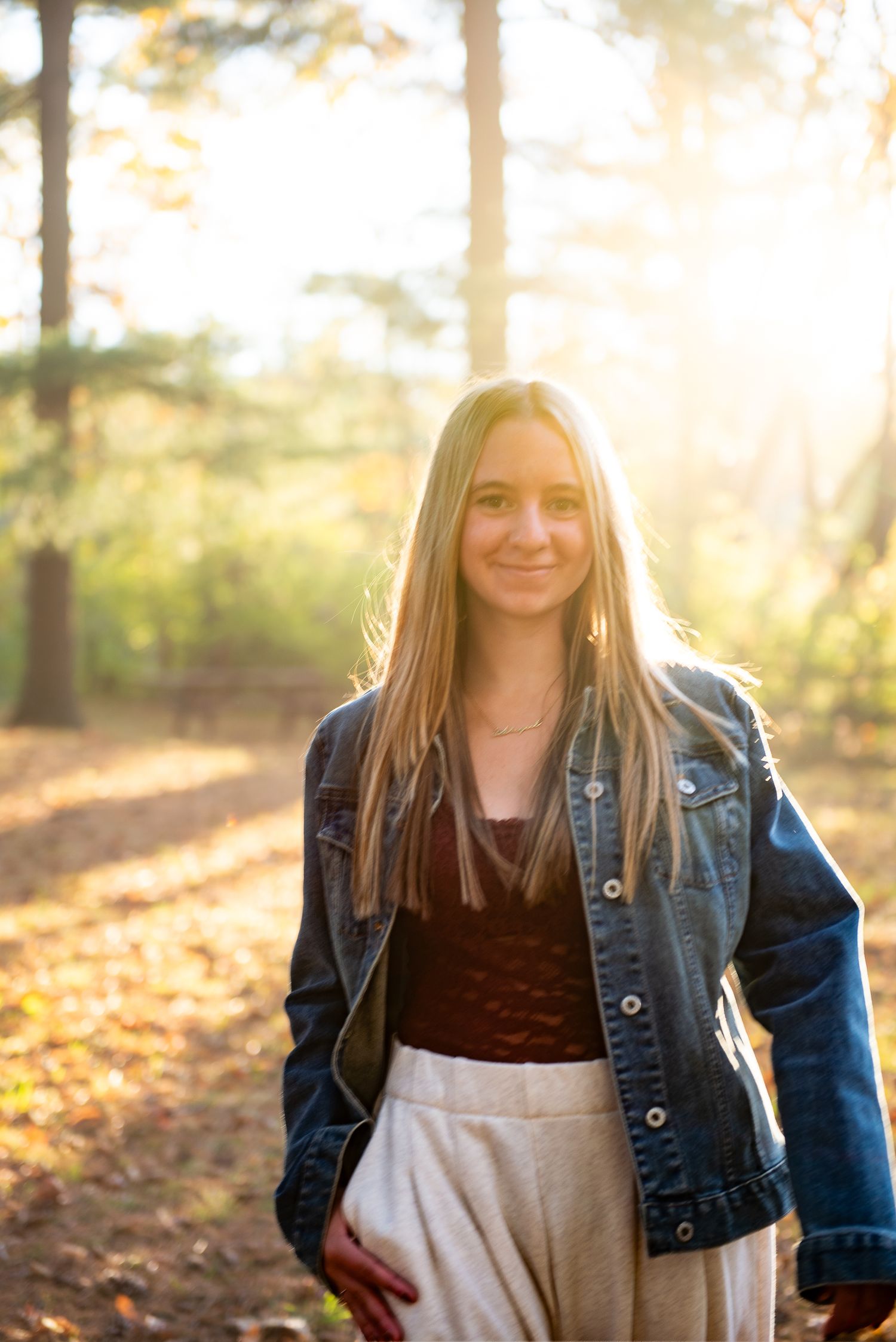 A person in a denim jacket stands in a sunlit forest during autumn with golden light filtering through trees.