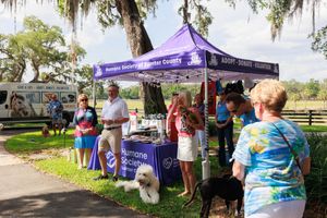 Visitors gather under purple tent canopies at an outdoor community event with dogs.
