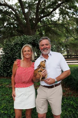 A small brown dachshund is held while posing outdoors under shady oak trees.