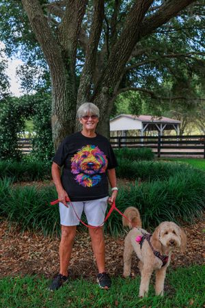 Person wearing colorful dog graphic t-shirt standing with two leashed dogs under large tree.