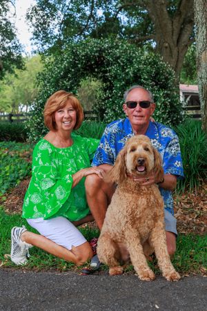 A large fluffy Goldendoodle dog poses outdoors with companions wearing bright green and blue shirts.
