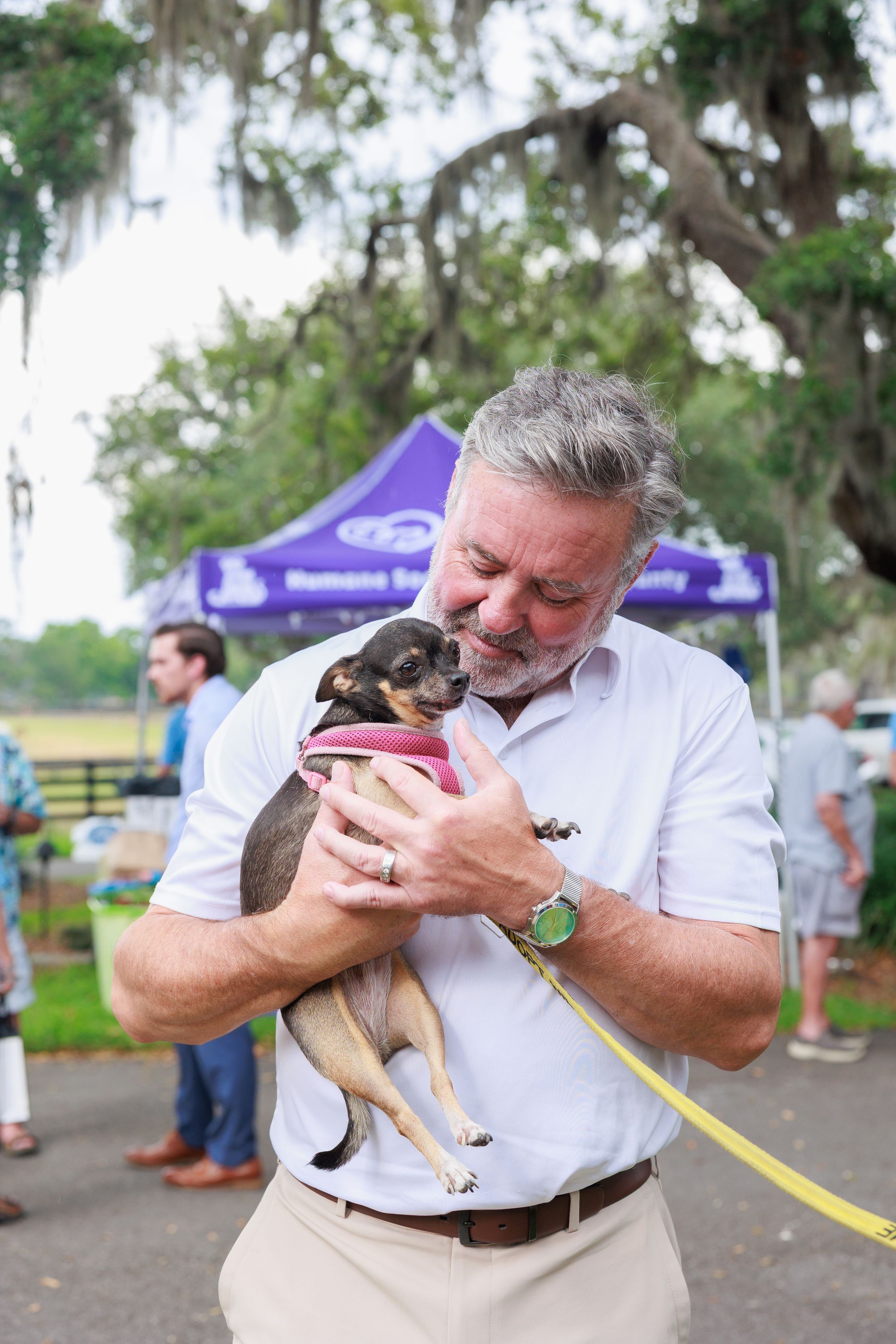 Person in white shirt lovingly holds and kisses small dog at outdoor community event with purple tent visible.