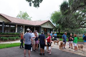 A group gathers outside a Spanish-style building with dogs on leashes at a community event.
