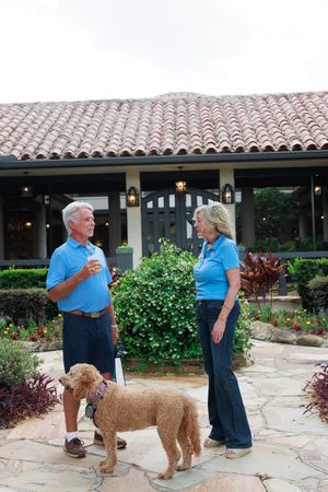 Two people in blue shirts interact with a large goldendoodle dog on a stone patio with Spanish-style architecture.