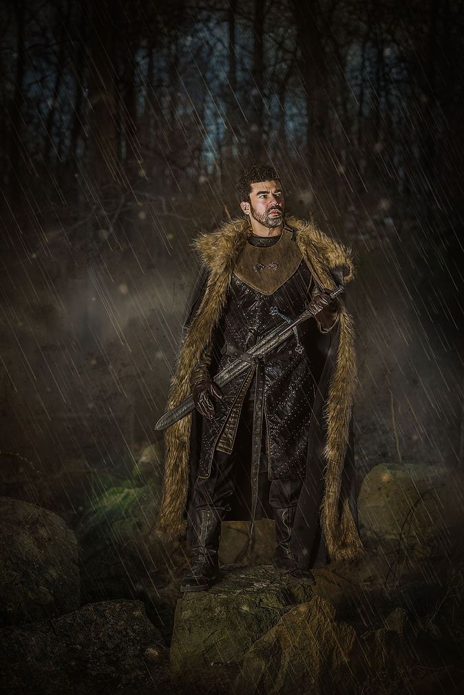 Medieval warrior man in fur cloak holding sword standing on rocks in dramatic rain scene.