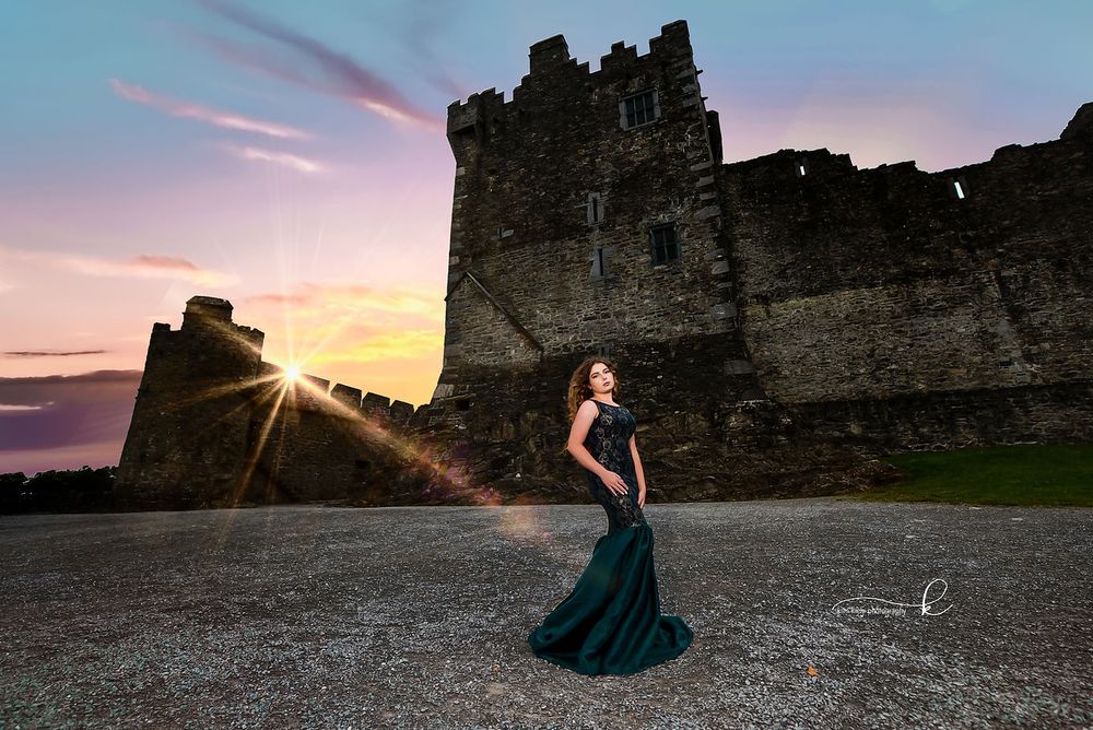 Woman in emerald green gown standing before historic castle at dramatic colorful sunset.