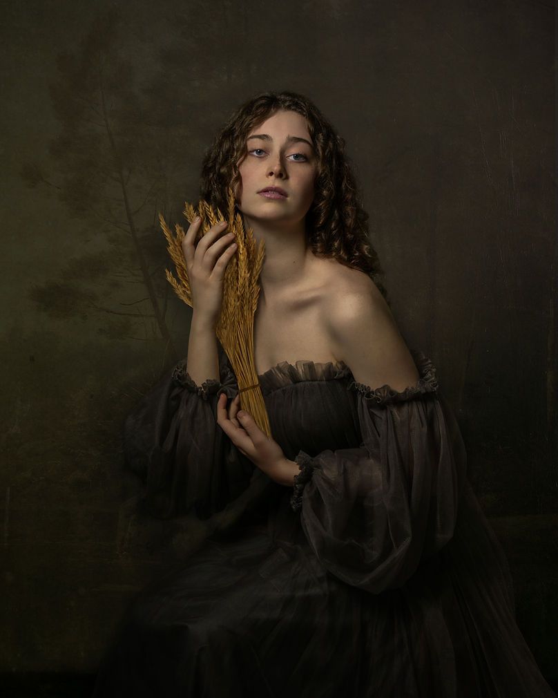 Young woman in grey off-shoulder gown holding golden wheat stalks in a dark classical portrait style.