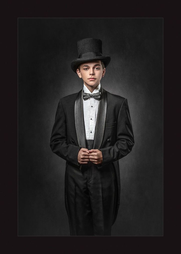 Young boy in formal black tailcoat tuxedo and top hat posing in a dramatic studio portrait.