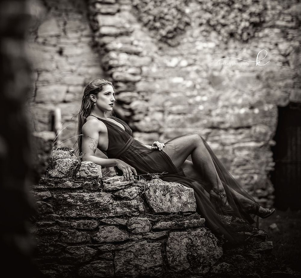 Black and white portrait of woman in elegant dress reclining on ancient stone ruins wall.