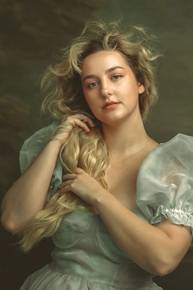 Young blonde woman in light blue puff-sleeve dress holding her hair in a classical portrait style.