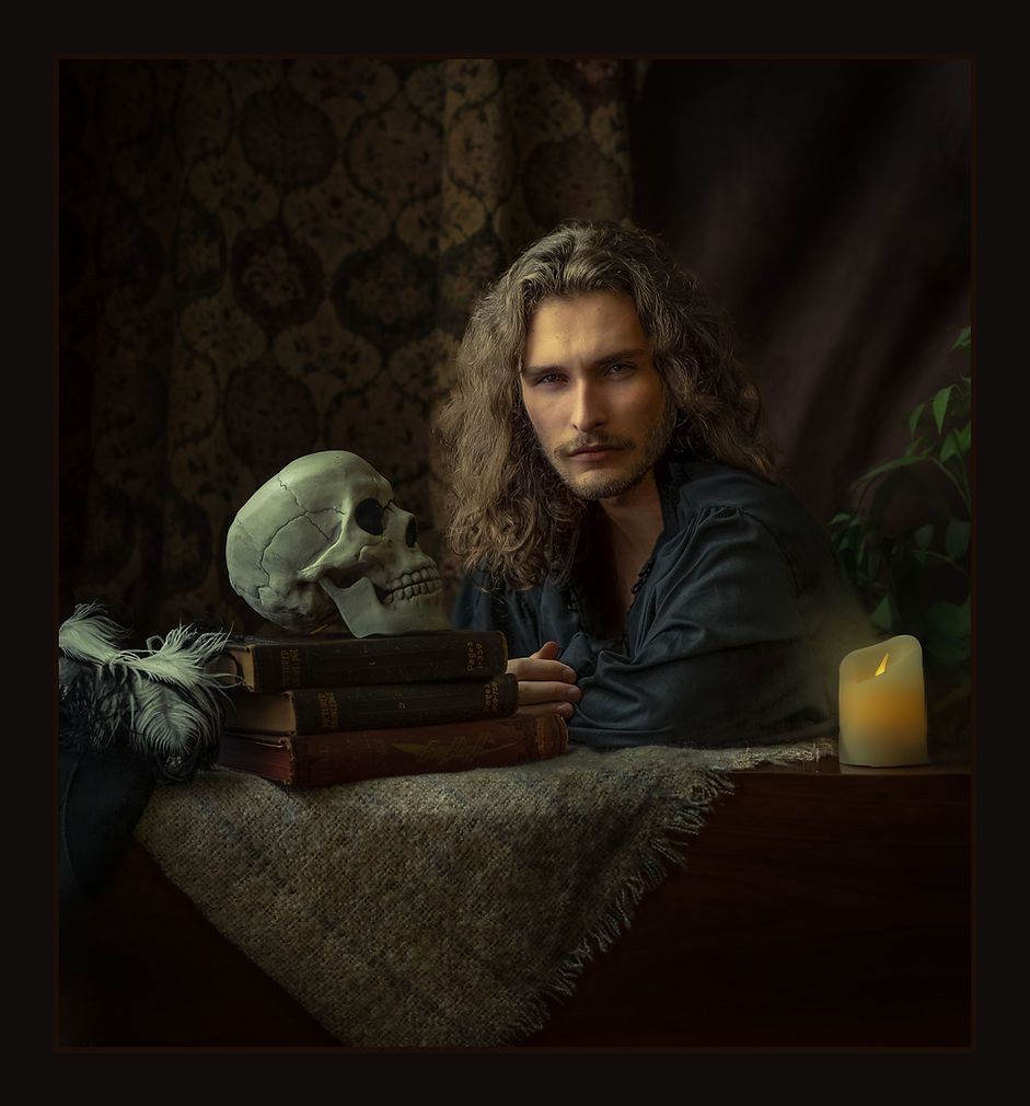 Long-haired man in dark shirt seated at table with skull, books and candle in baroque style.