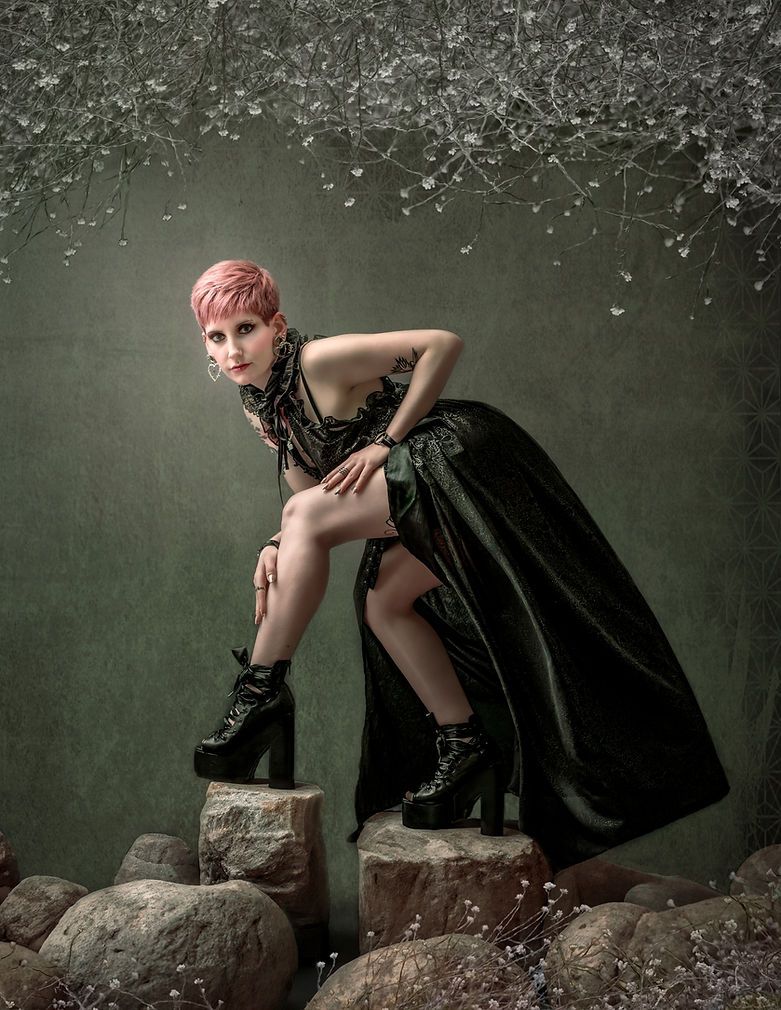Woman in dark green gown and black platform boots poses dramatically on rocks in an artistic fantasy portrait.