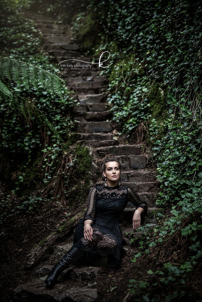 Woman in black lace dress seated on moss-covered stone steps surrounded by ferns and ivy.