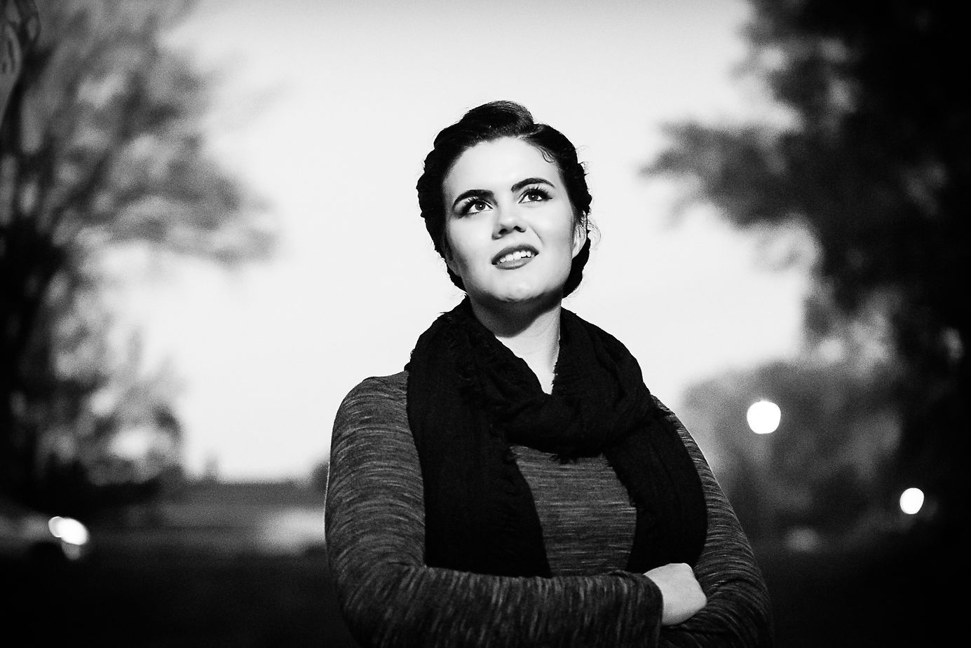 Black and white portrait of a young woman with a scarf looking up outdoors.