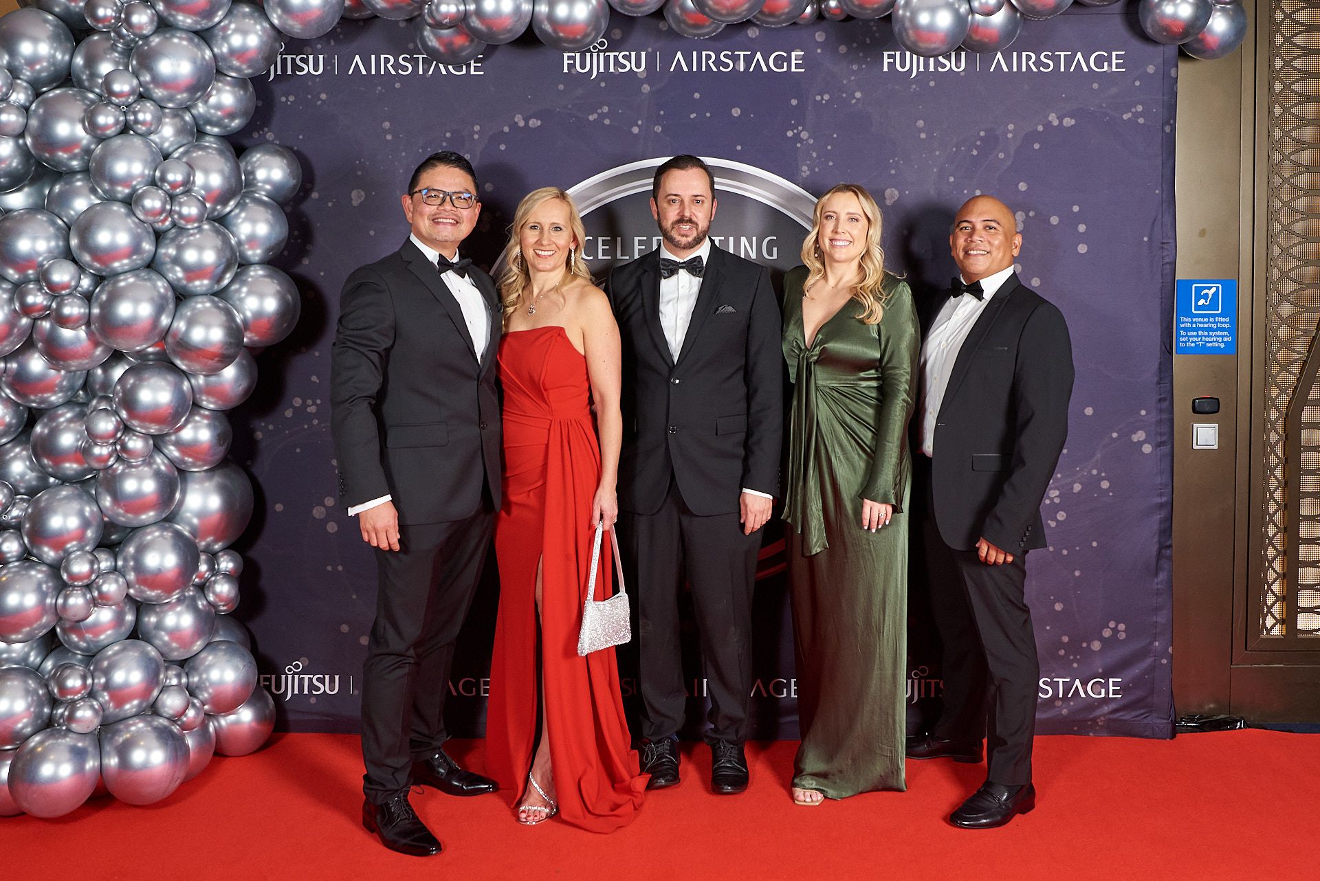 Group of people posing for photos on red carpet with silver balloon arch decorations at formal event.