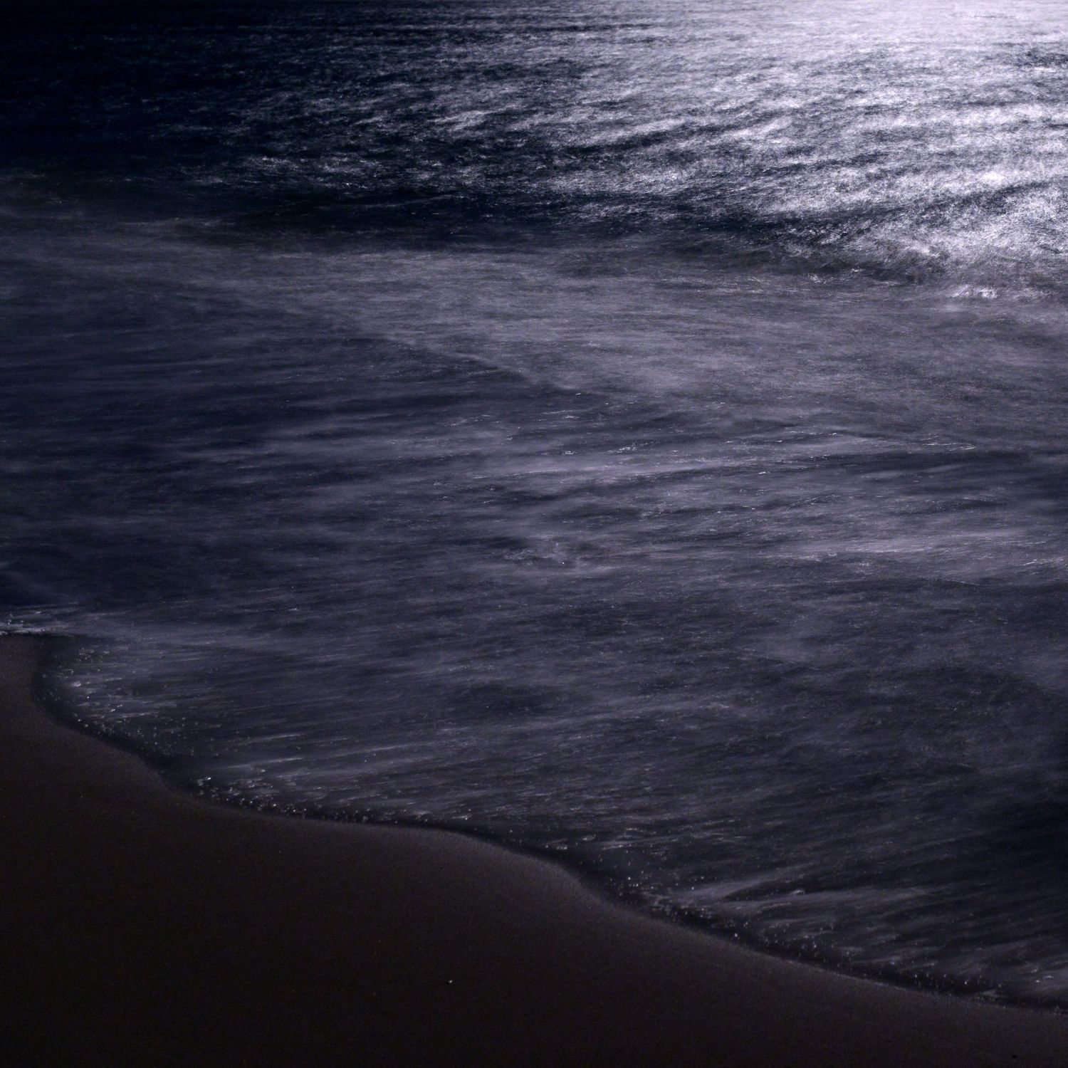 Waves gently rolling onto a dark sandy beach at night with moonlight reflecting off the rippling water surface.