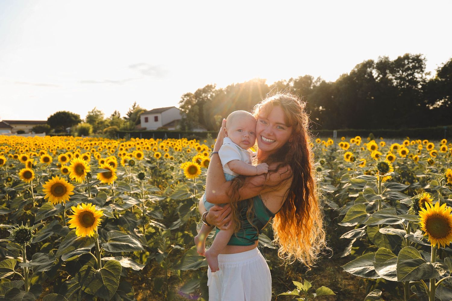 Joyful mother holding baby in golden sunflower field at sunset with warm backlight.