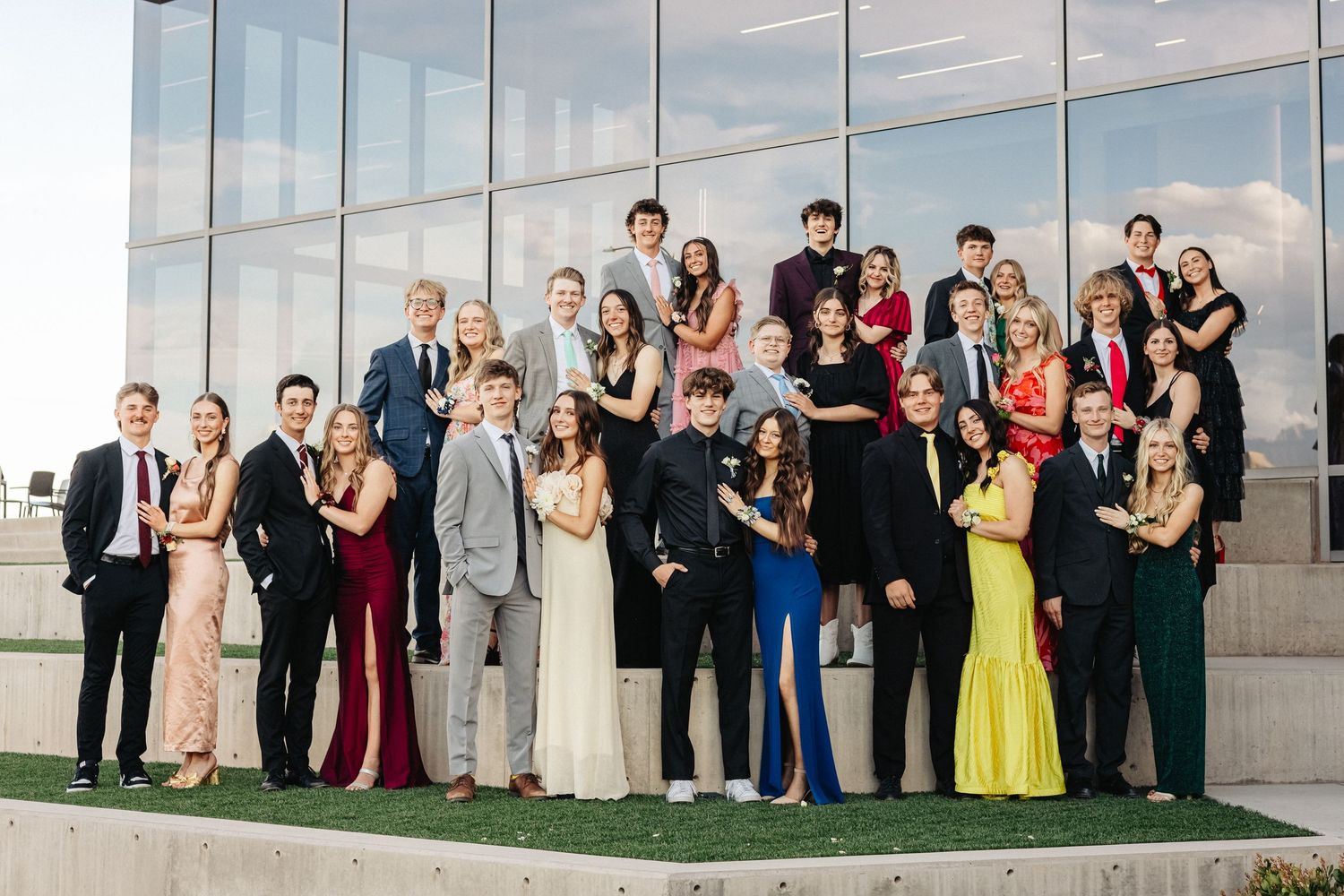 A formal group photo of people dressed in suits and gowns standing together in front of a glass building exterior.