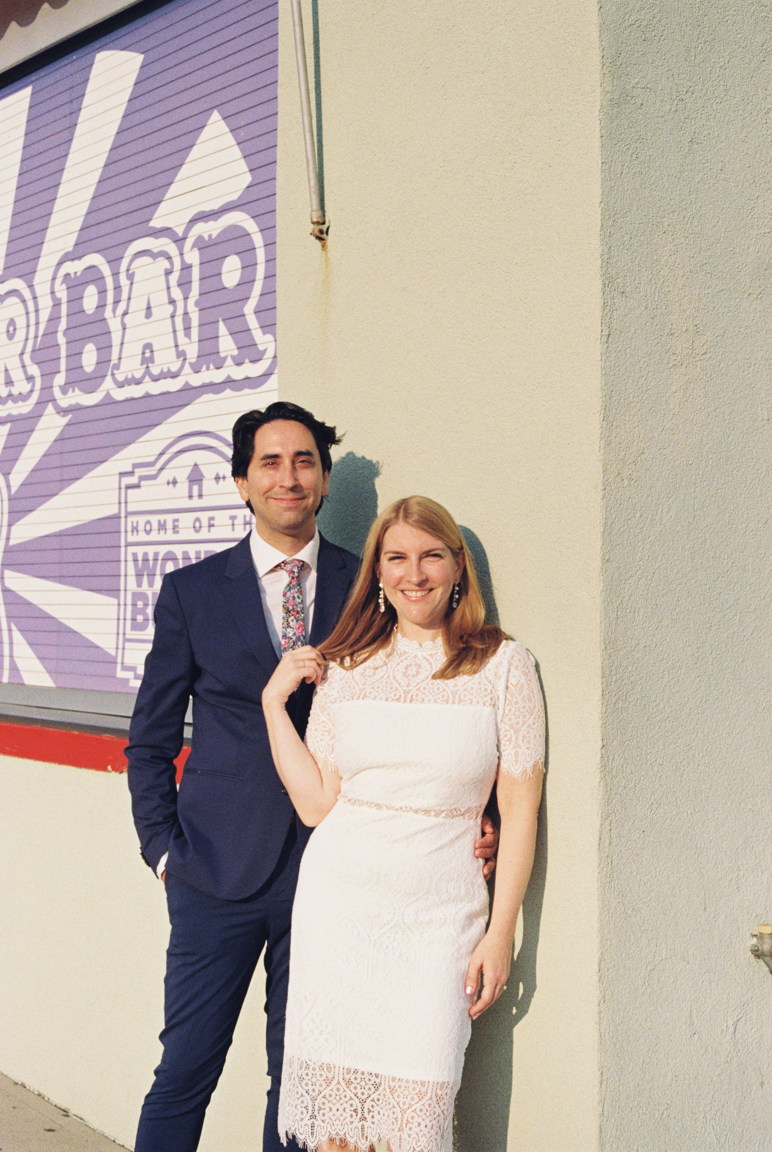 A couple poses against a purple wall with signage in an urban setting.