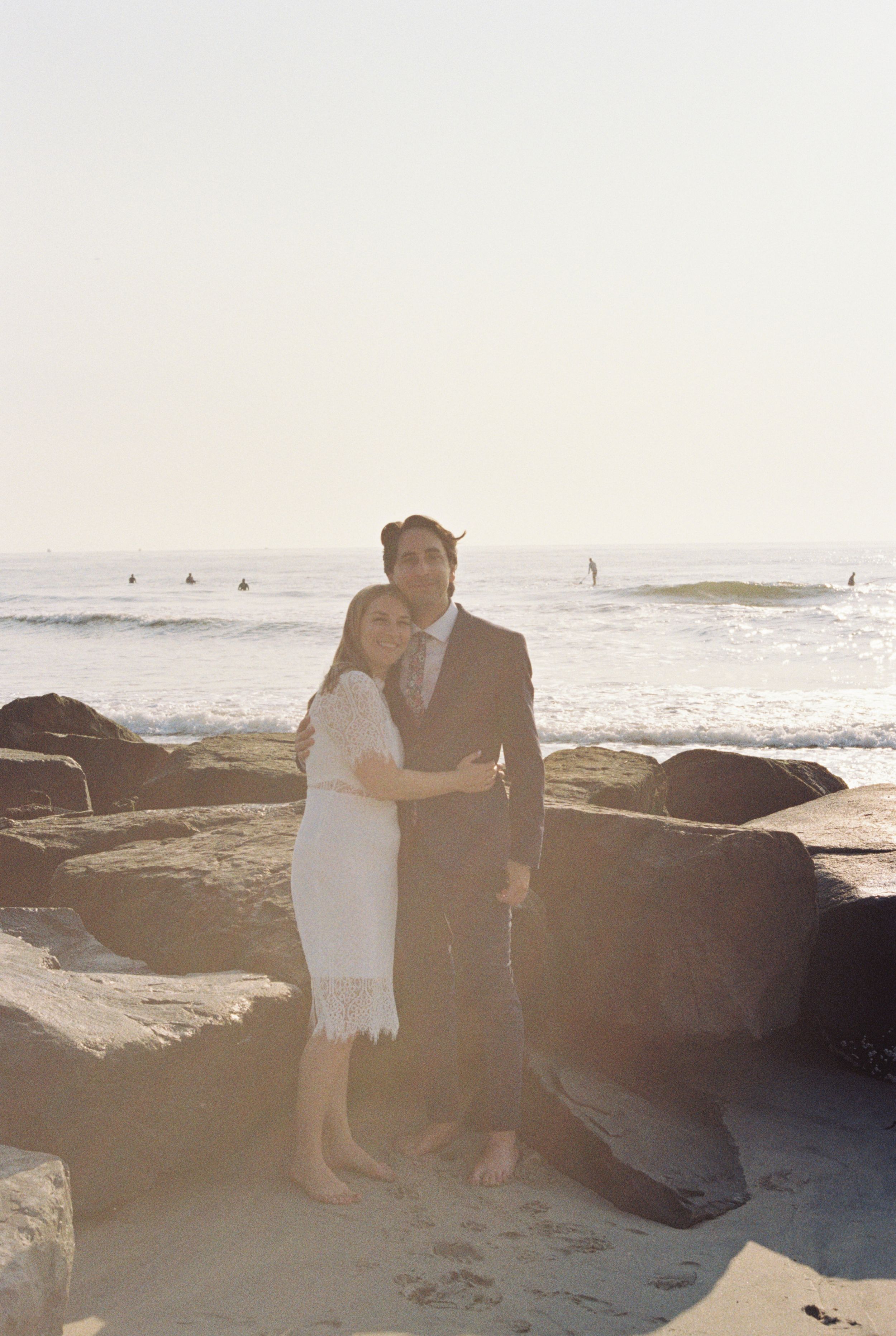 A couple embraces on rocky beach cliffs at sunset with ocean waves in background.