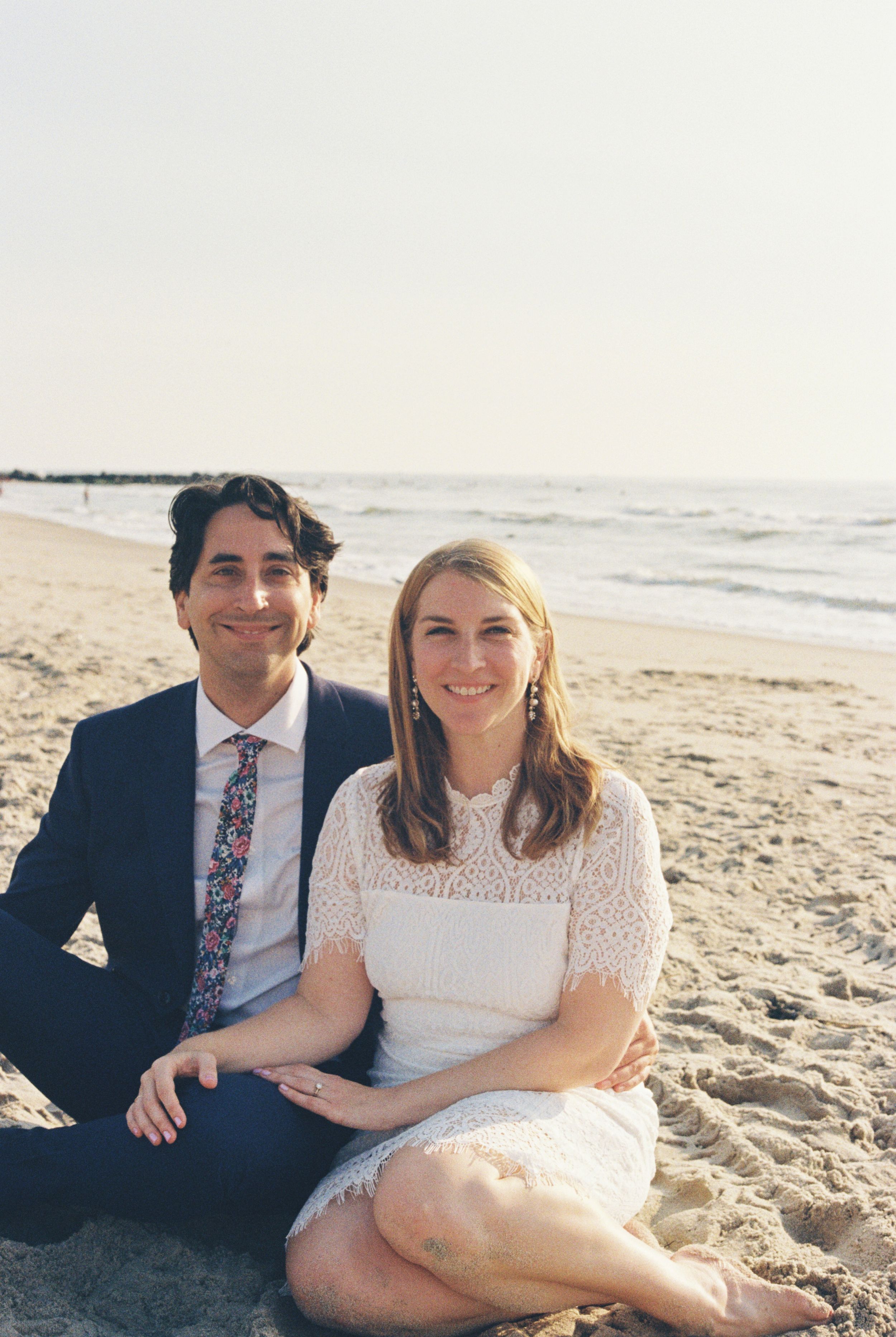 A couple sits together on a sandy beach in a series of romantic engagement photos.