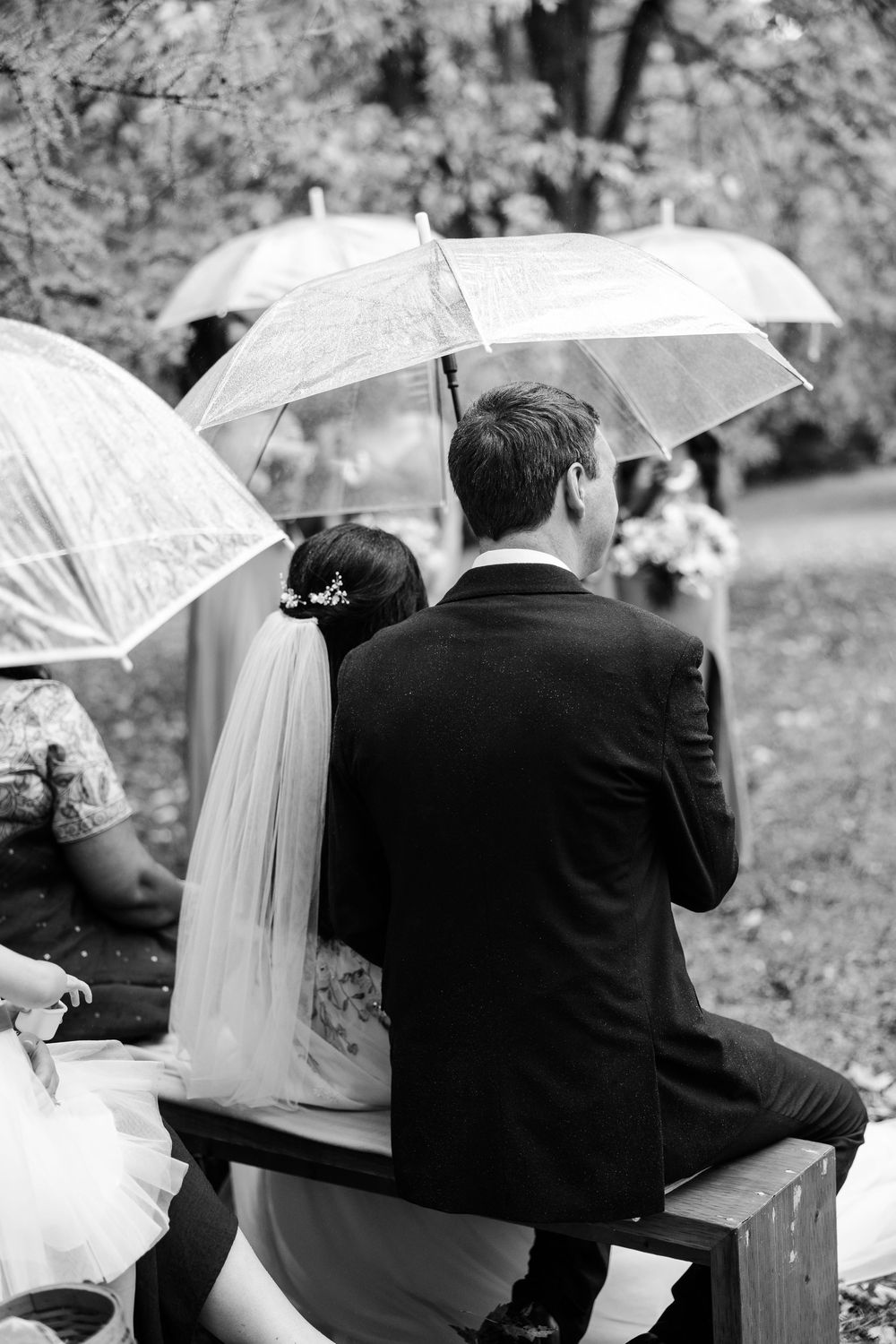 A black and white wedding photo series showing a couple sitting together under a white umbrella in a garden setting.