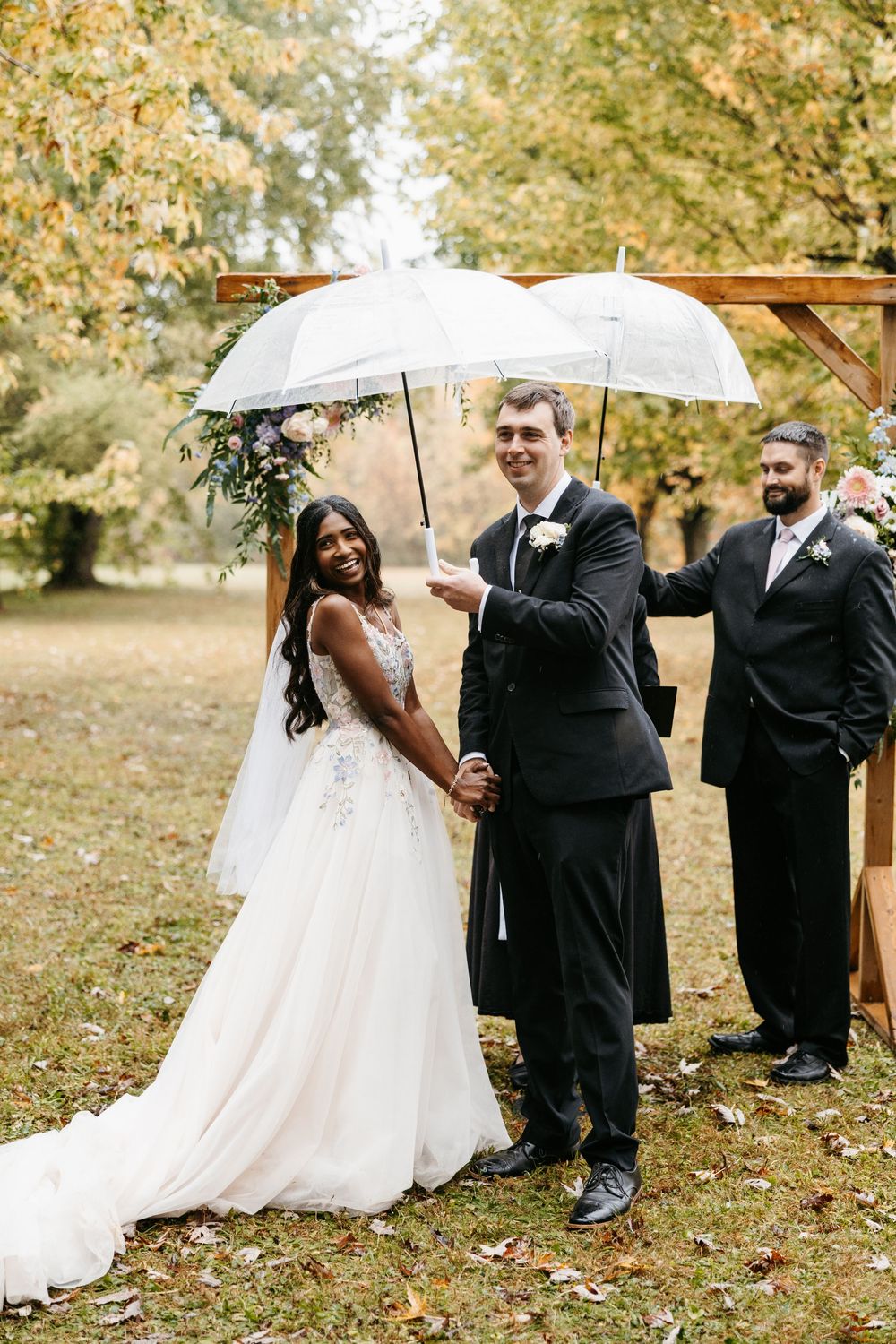 Wedding party poses with white umbrellas under autumn trees during outdoor fall ceremony.