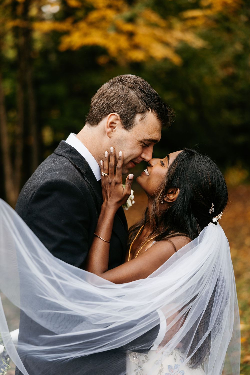 A romantic wedding moment captured outdoors with flowing white veil and autumn foliage in the background.