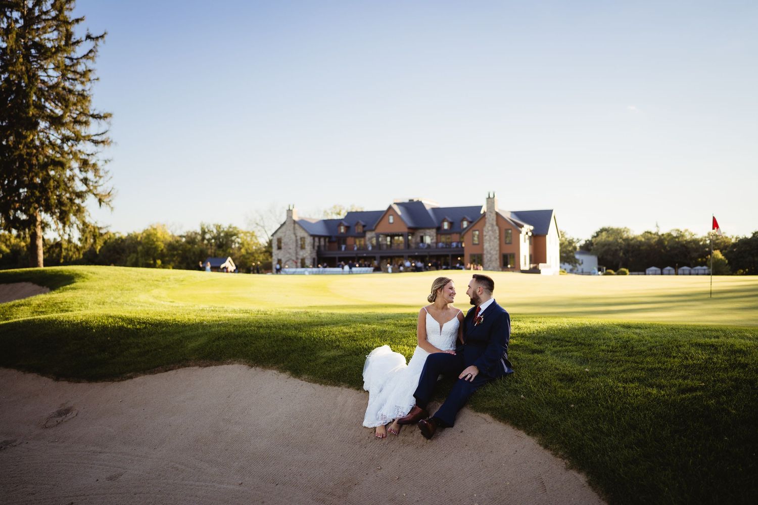 A couple sits together on a grassy hillside during golden hour at sunset.
