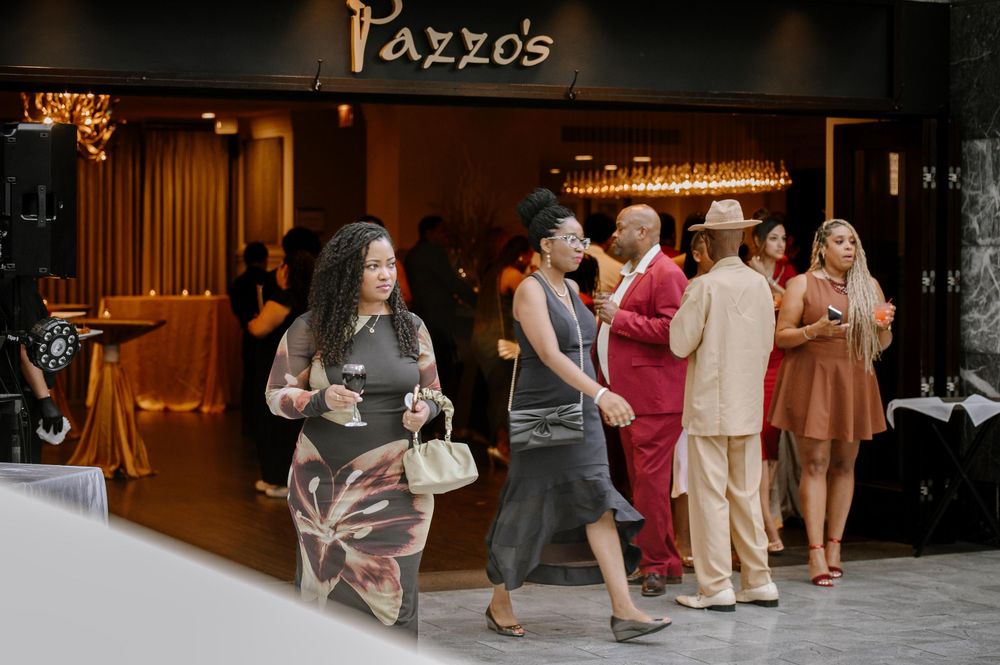 A line of wedding guests dancing in a dimly lit reception venue with Pazzo's signage visible.