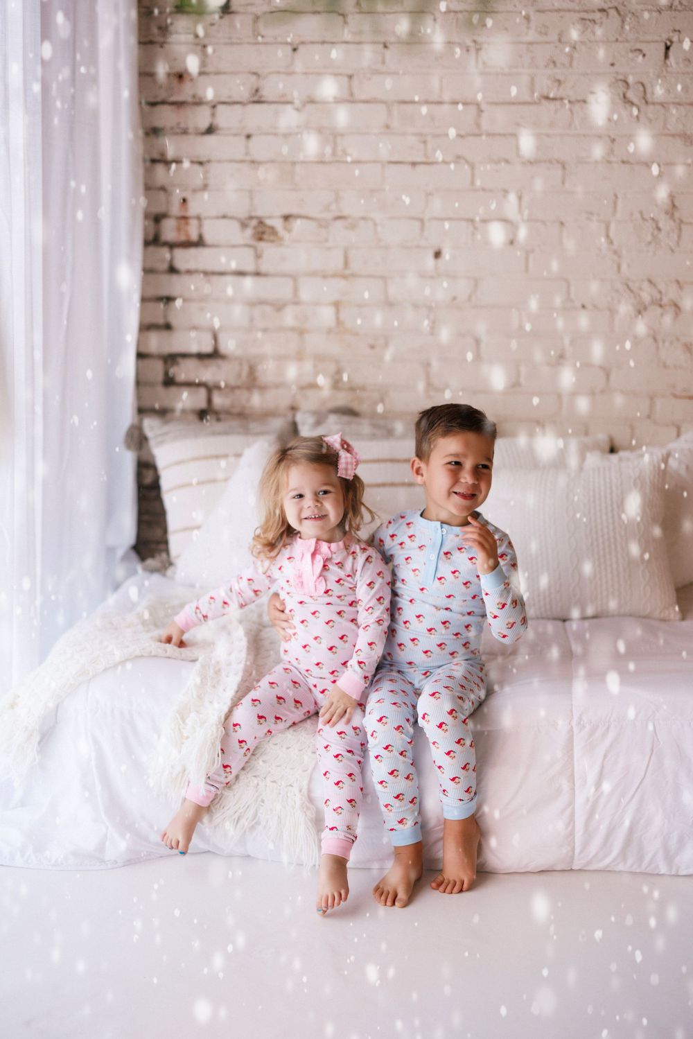 Children in matching heart-print pajamas sit and play on a white couch against a rustic brick wall background.