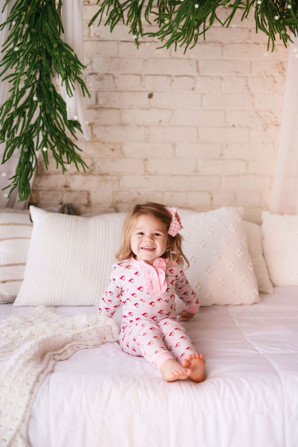 Adorable child in red and white patterned pajamas laying on white bedding with greenery decor above.