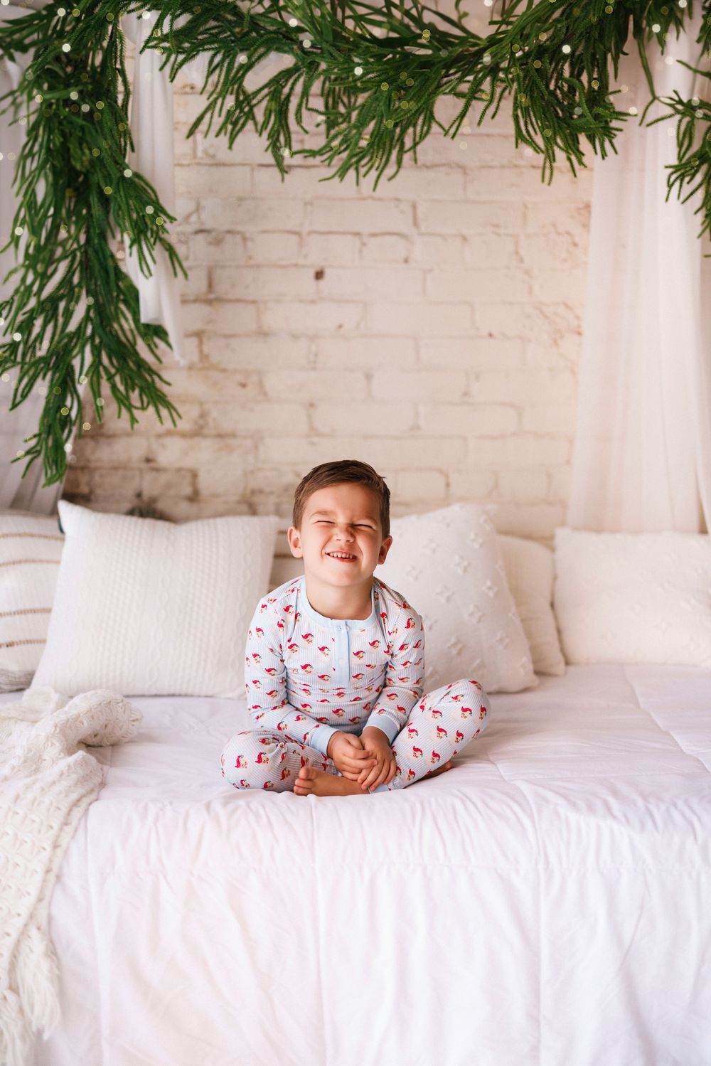 Child in festive sleepwear playing and smiling on white bed with evergreen garland decoration.