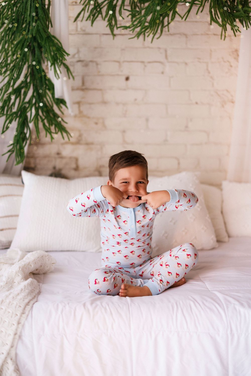 Playful scene of child in holiday pajamas lounging on white bedding with greenery overhead.