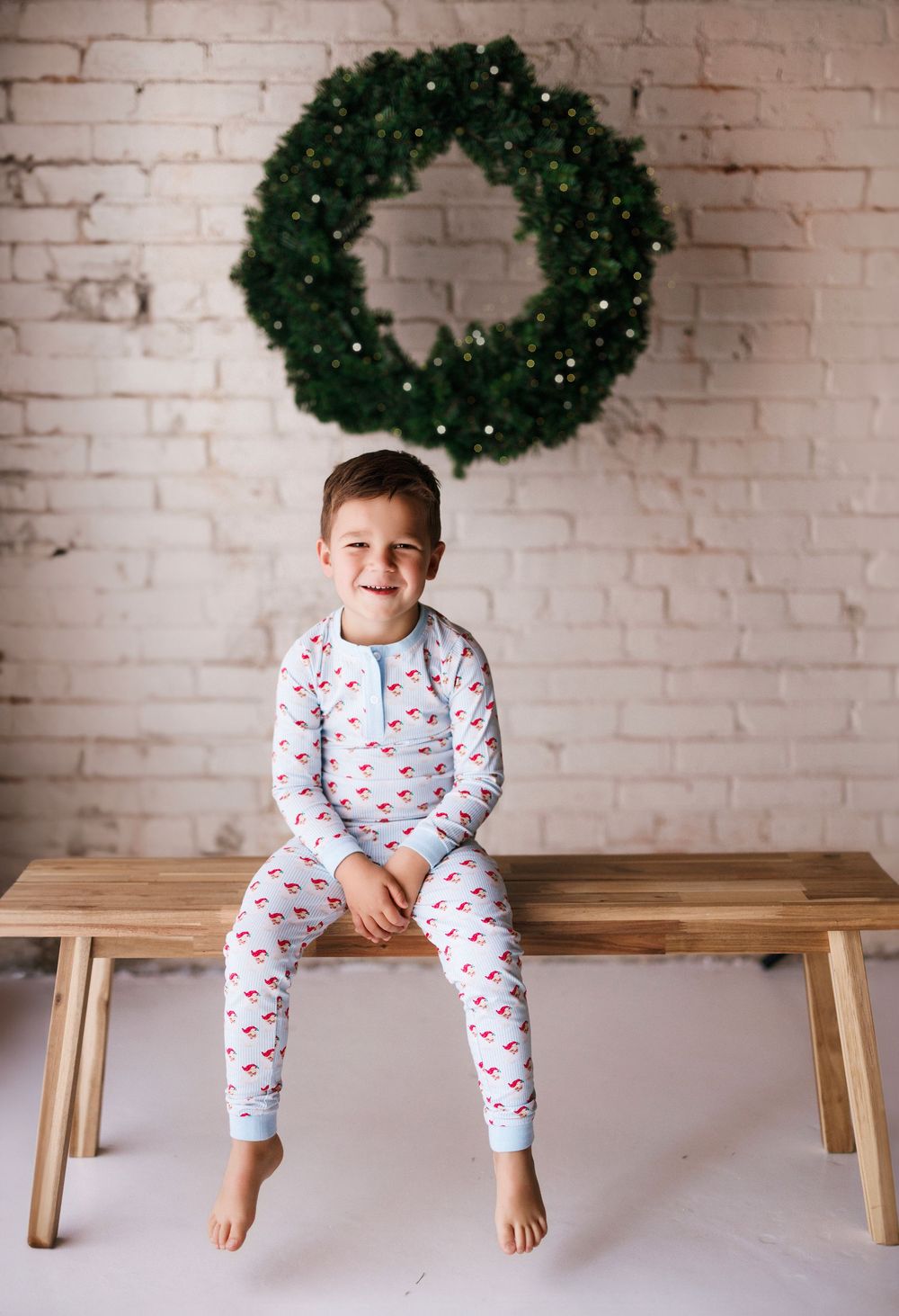A child in holiday pajamas sits on a wooden bench beneath a green Christmas wreath on a white brick wall.