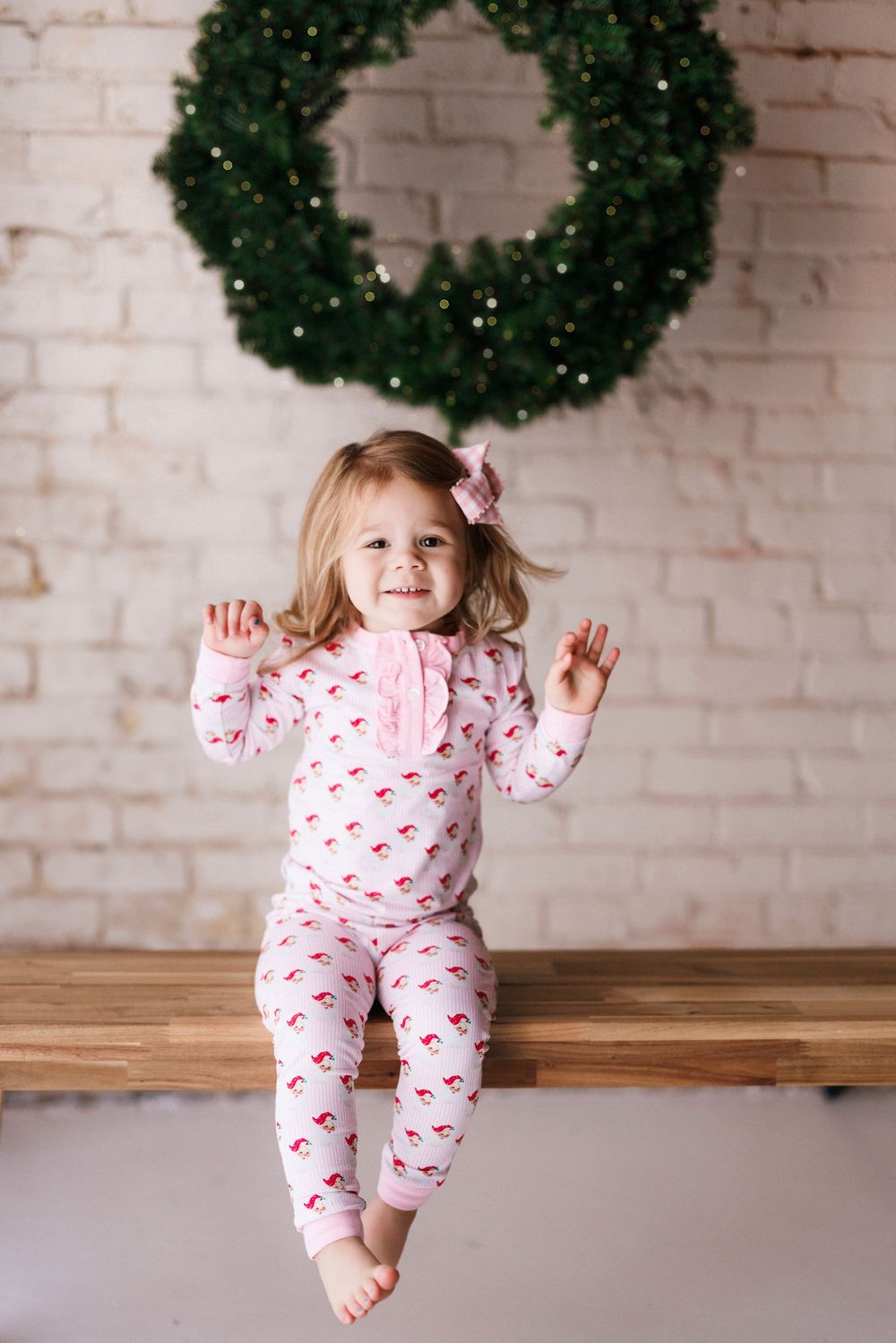 A child in heart-patterned pajamas plays energetically on a wooden bench against a white brick wall with wreaths.