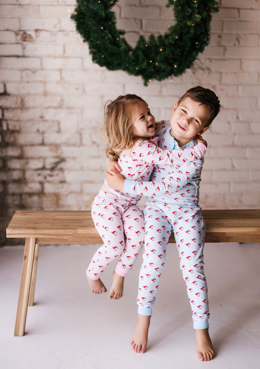 Two children in matching Christmas pajamas playing together on wooden bench under wreath decoration.
