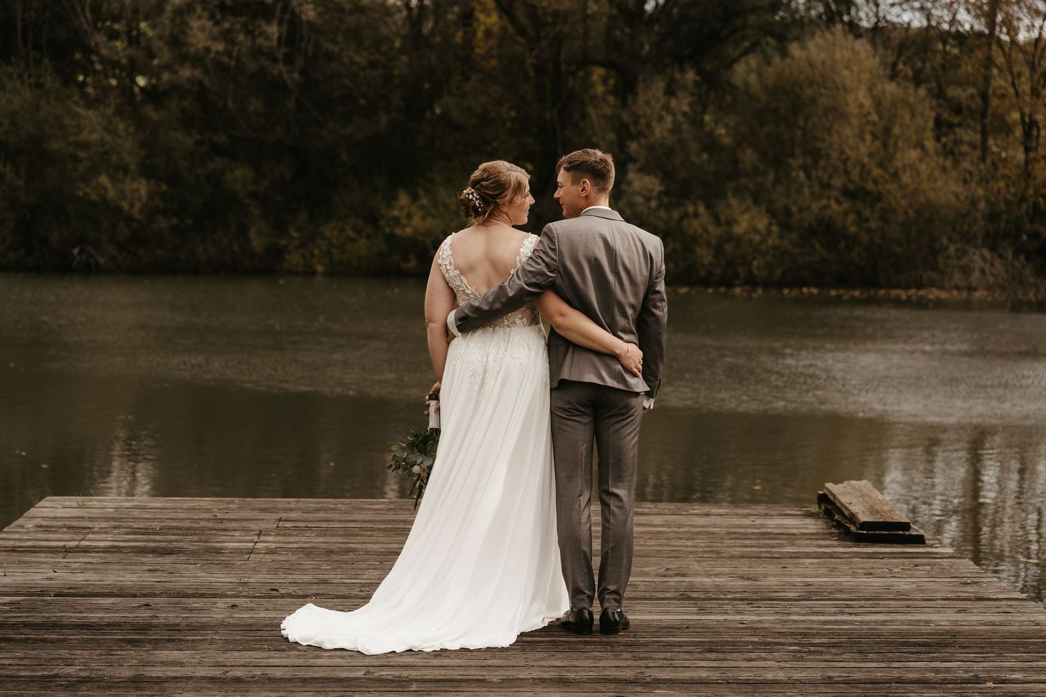 A couple in wedding attire stands together on a wooden dock overlooking a serene lake during autumn.