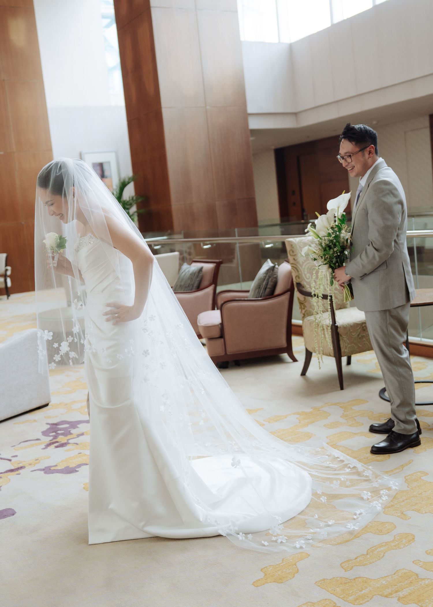 Groom in gray suit smiles holding white bouquet during first look moment with bride in wedding venue.