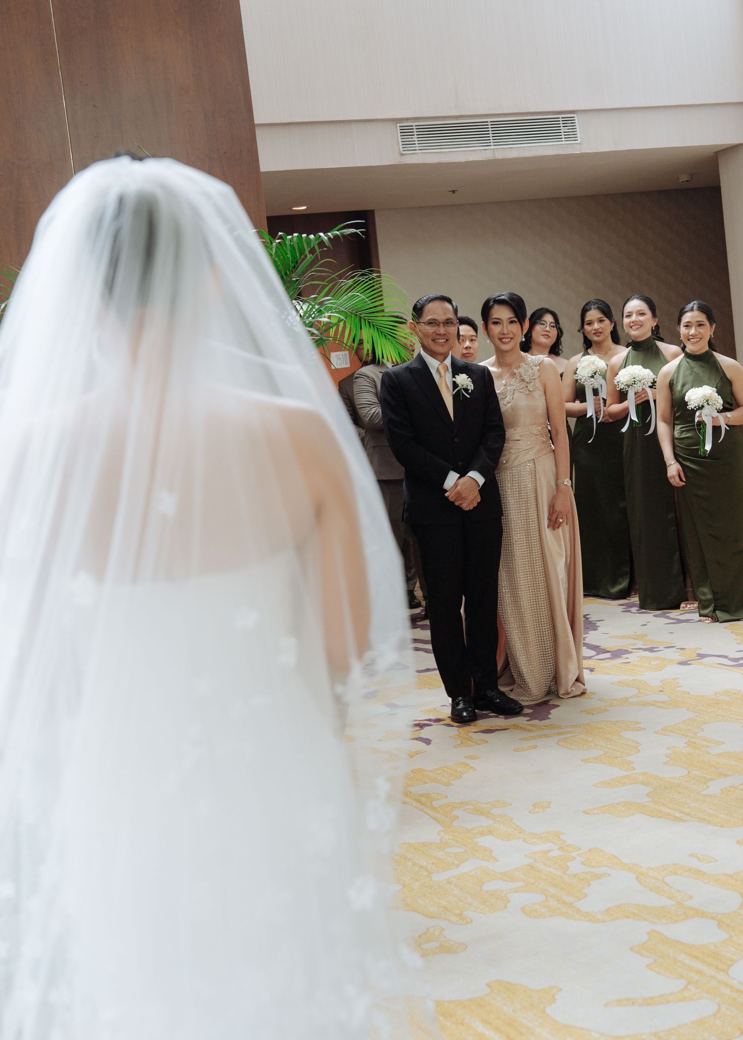 Bride in white veil approaches smiling parents and bridesmaids in olive green dresses at wedding venue.