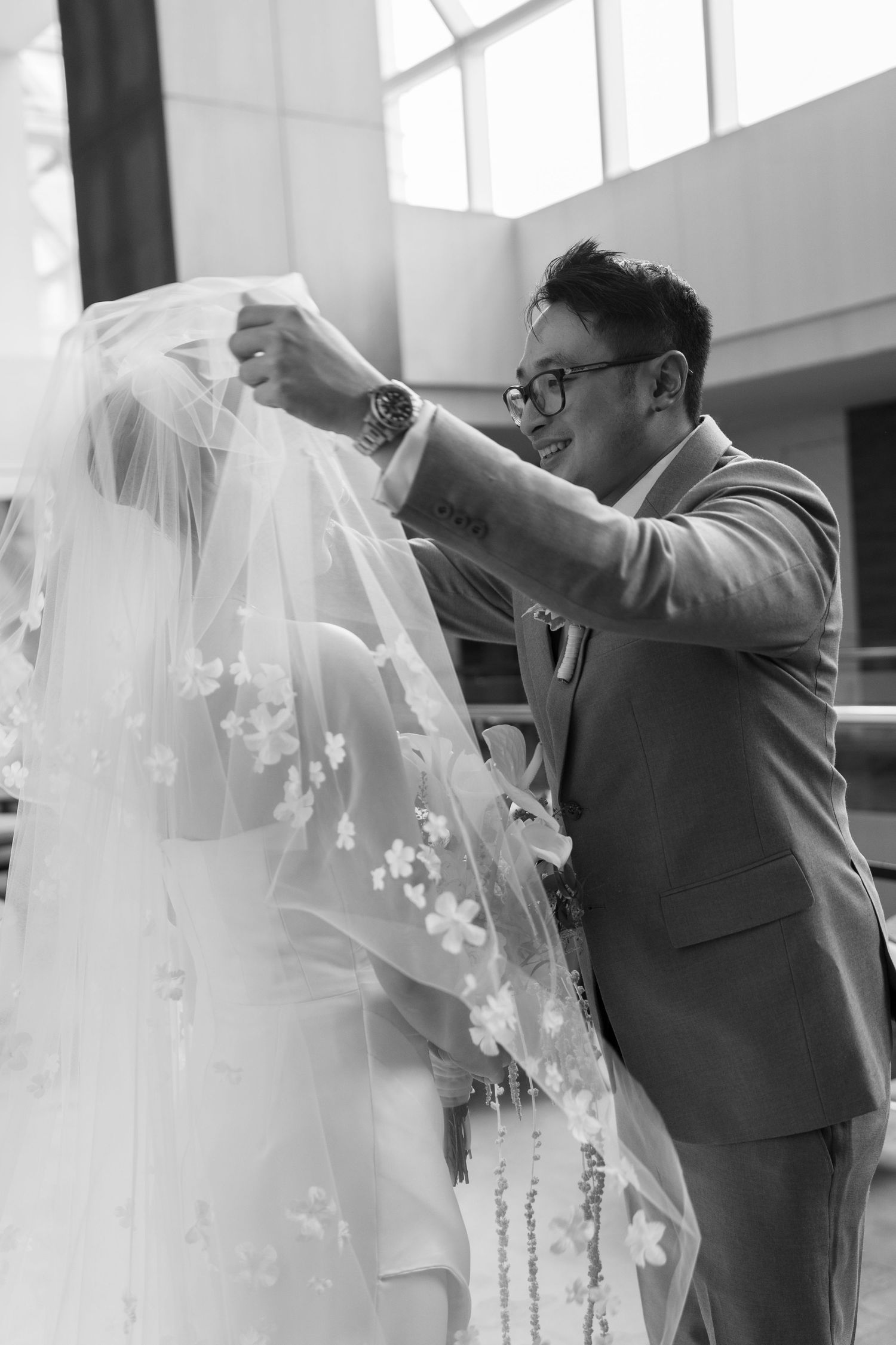 Groom in gray suit lifts bride's floral cathedral veil in black and white wedding ceremony photo.