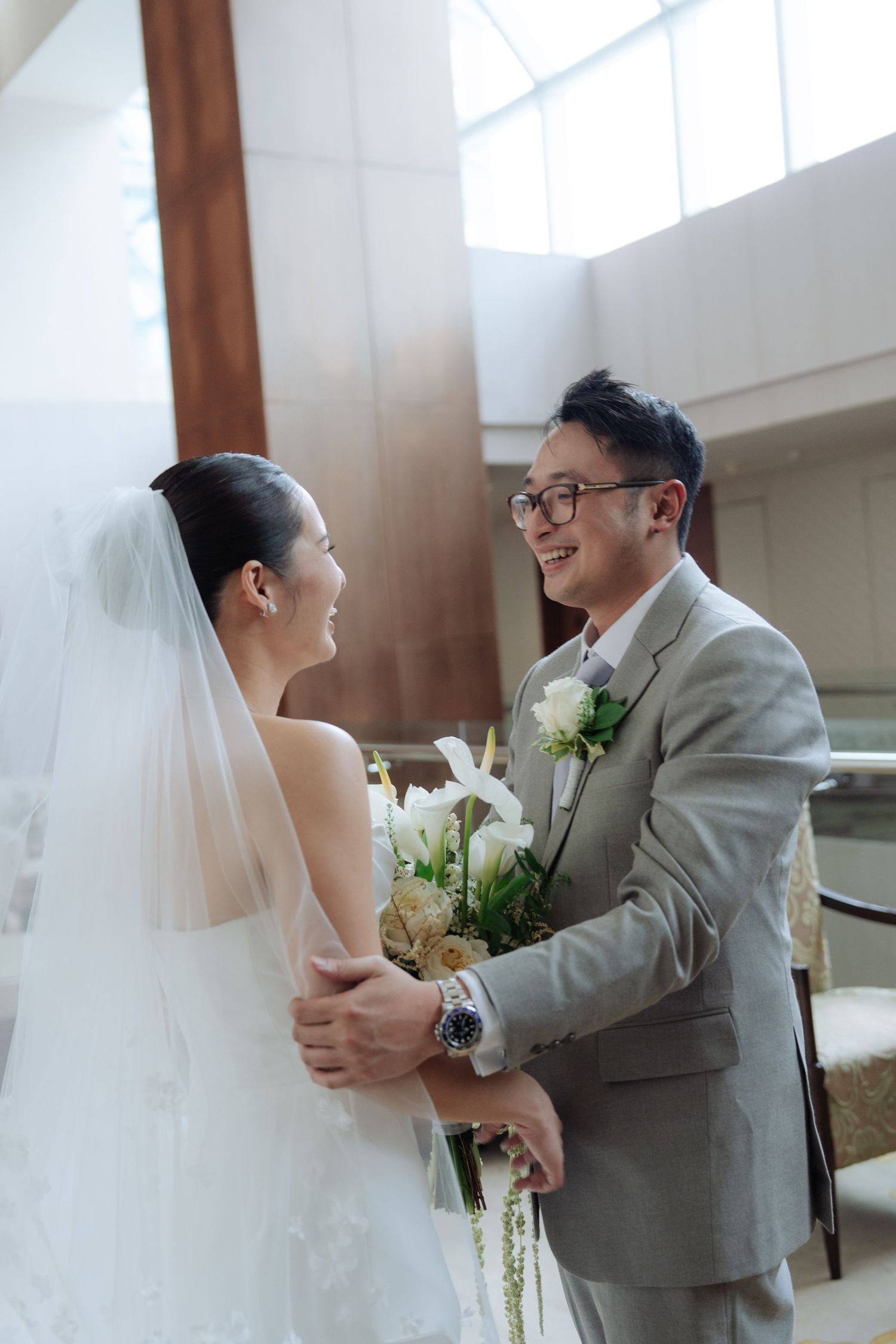 Smiling bride and groom share joyful first look moment, groom holding white floral bouquet in gray suit.