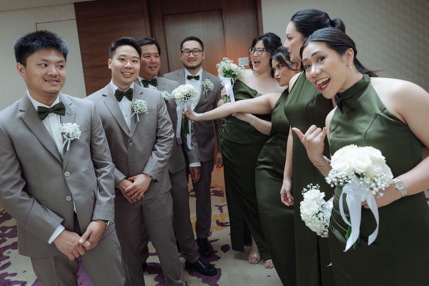 Bridesmaids in olive dresses playfully interact with groomsmen in gray suits at indoor wedding reception.