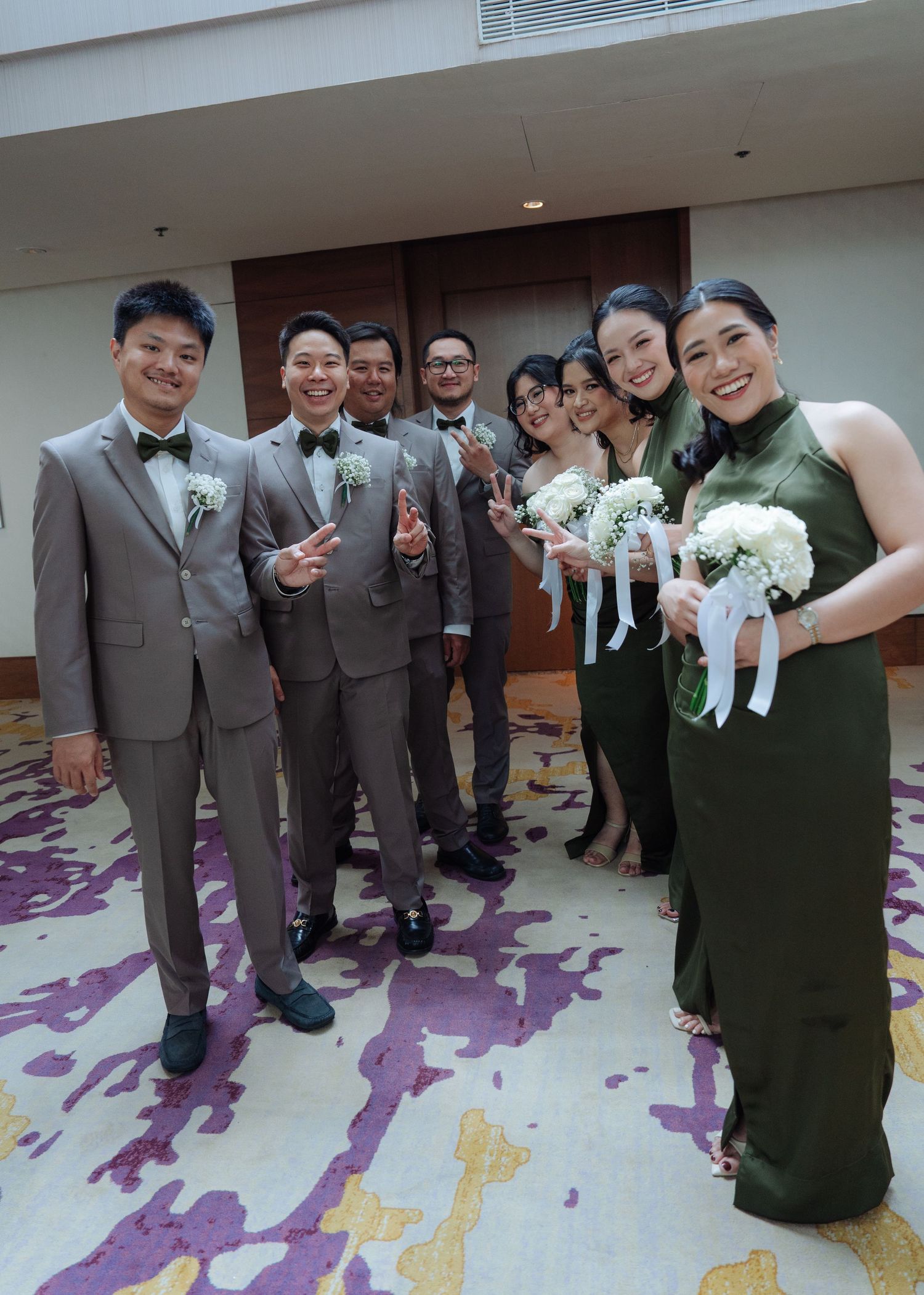 Wedding party group photo with groomsmen in gray suits and bridesmaids in olive dresses holding white bouquets.