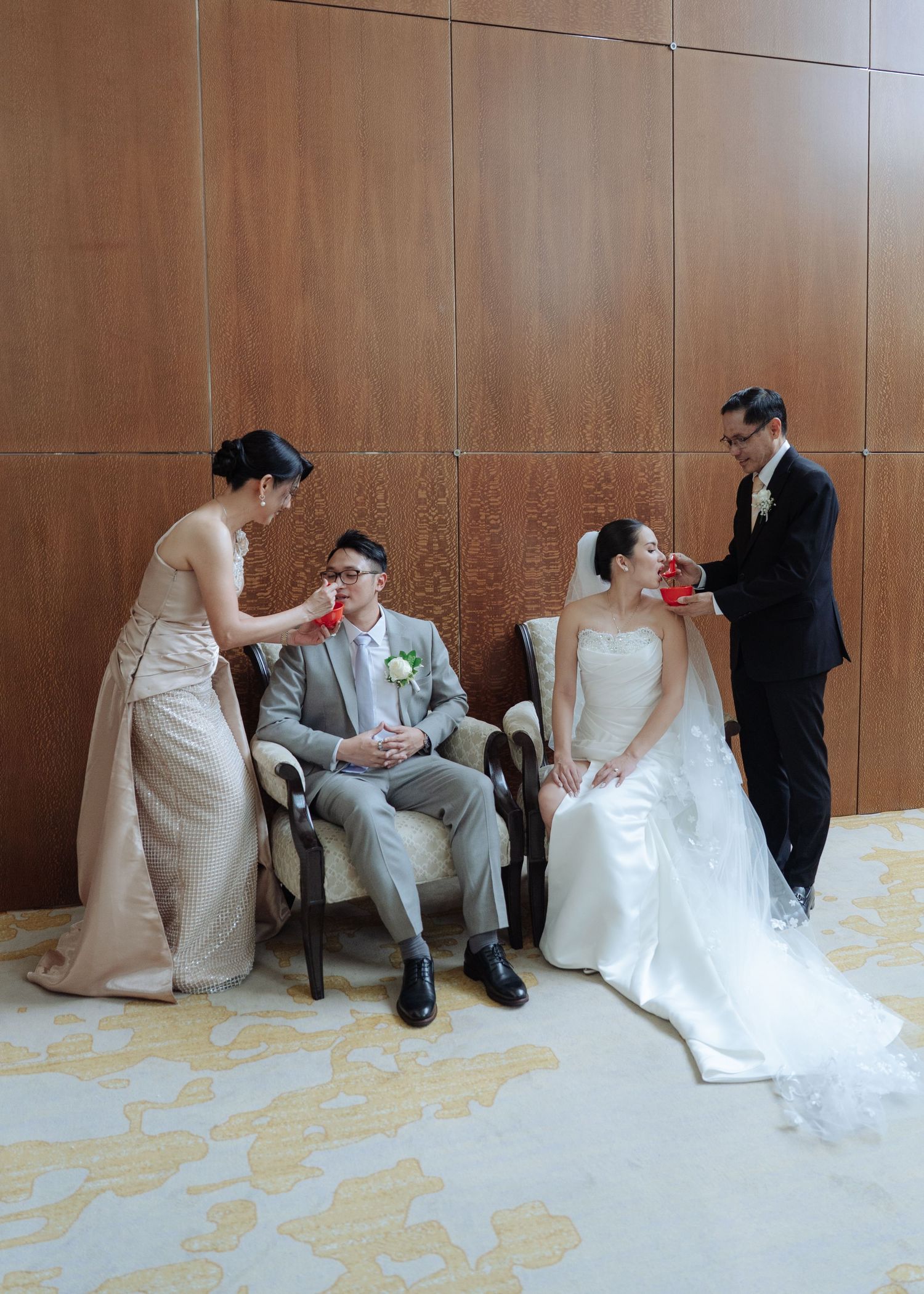 Bride and groom seated in chairs while attendants serve them drinks during a traditional Chinese wedding tea ceremony.