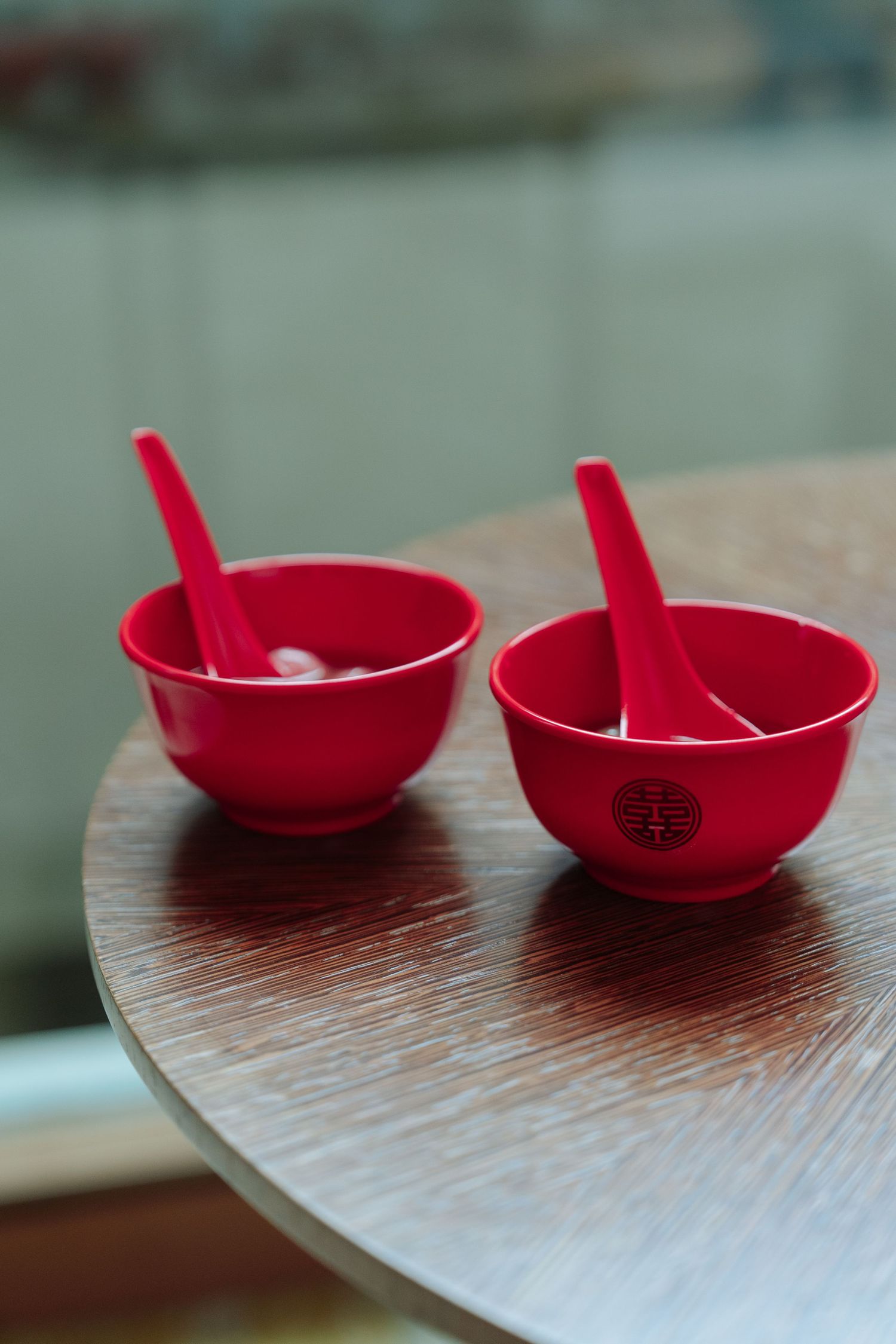 Two red Chinese wedding bowls with spoons featuring double happiness symbol on a wooden table.