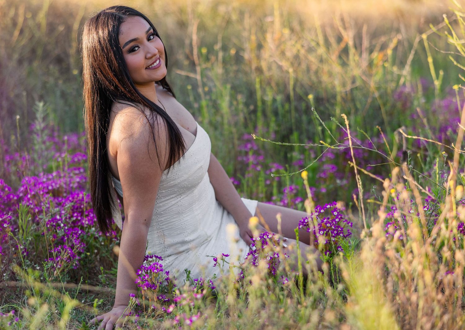 A woman in a white dress poses gracefully in a field of purple wildflowers during golden hour.