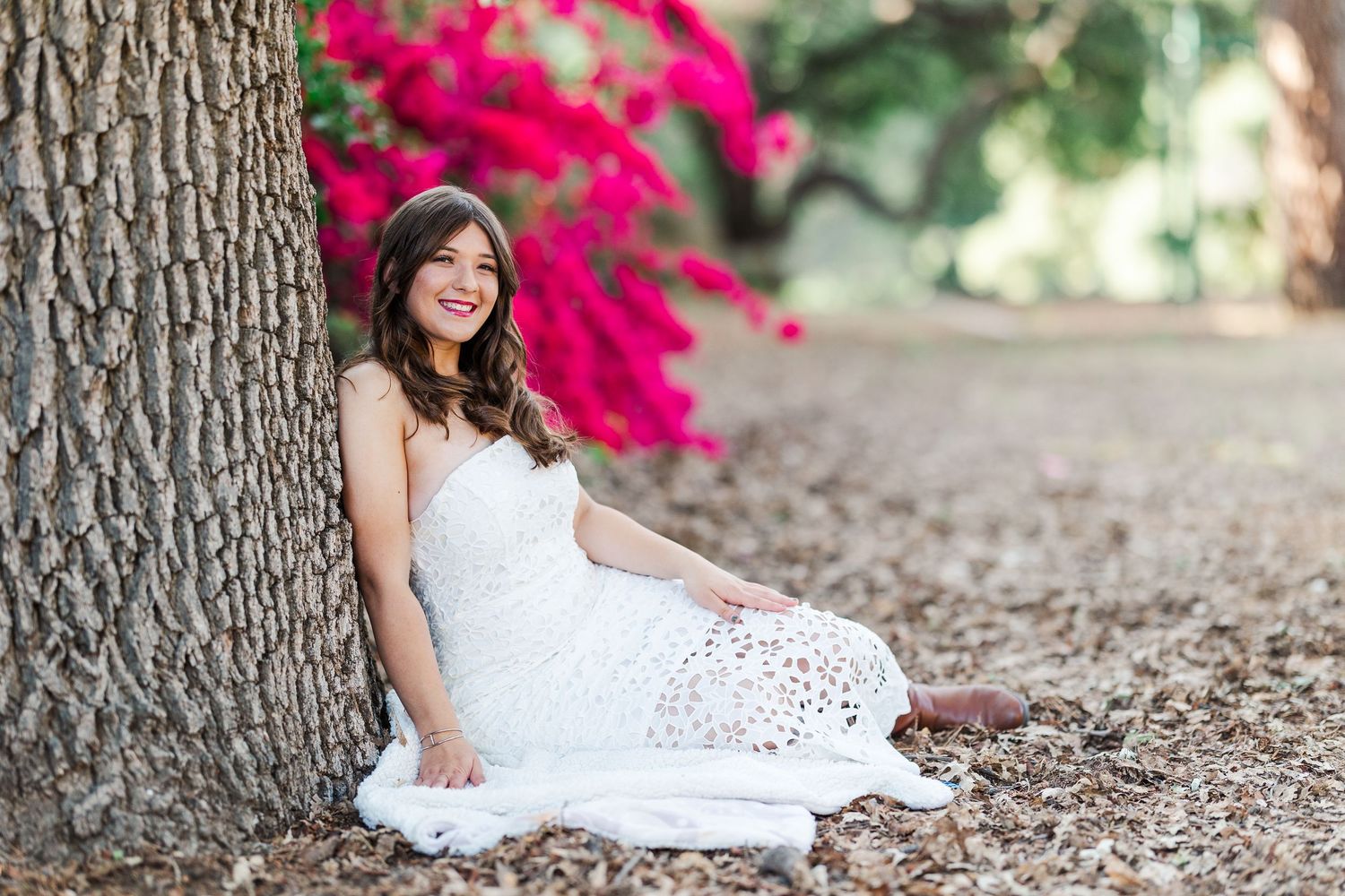 A person in a white lace dress poses elegantly against a backdrop of vibrant pink bougainvillea flowers and trees.