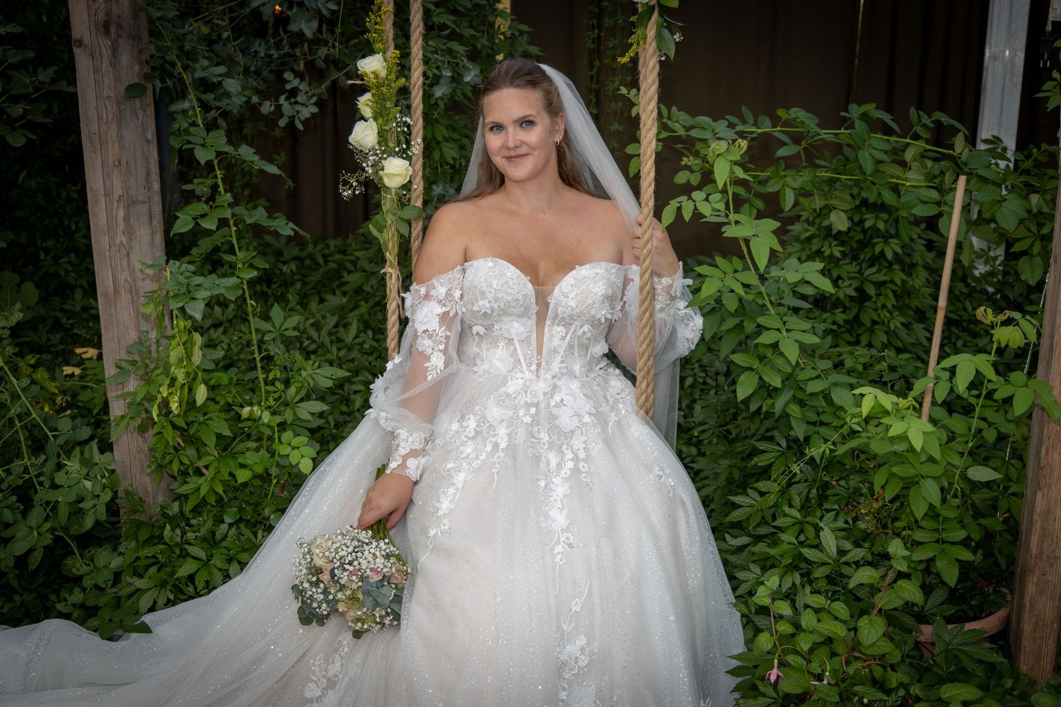 Bride in stunning off-shoulder lace ball gown with veil sitting on a garden swing surrounded by greenery.